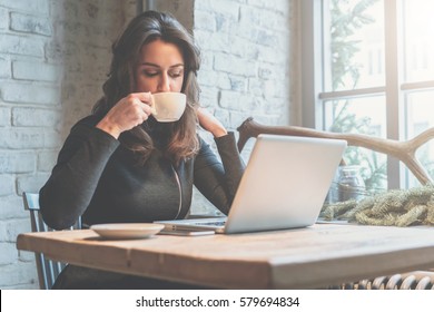 Young businesswoman sitting in coffee shop at  table in front of laptop and drinking coffee. On background white brick wall and window. Girl shopping online, blogging, checking email.Online education.