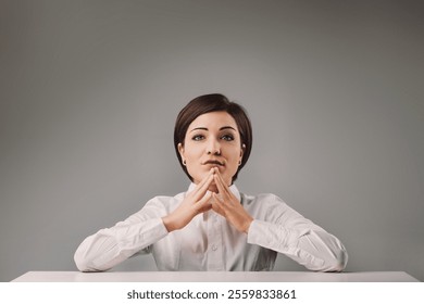 Young businesswoman sitting at a desk with her hands clasped, thinking and focused, exuding confidence and professionalism in a studio setting with a neutral background - Powered by Shutterstock