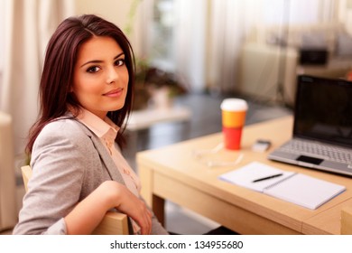 Young Businesswoman Sitting At Desk And Working. Smiling And Looking Back At Camera