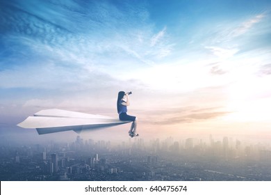 Young businesswoman sitting on a paper aeroplane while flying above a city and looking through a binoculars
