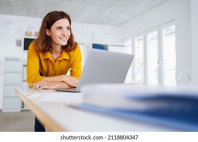 Young businesswoman smiling to herself in happy anticipation of the fulfillment of her dreams and ambitions as she leans over a desk at her laptop computer in a low angle view