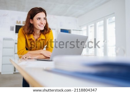 Young businesswoman smiling to herself in happy anticipation of the fulfillment of her dreams and ambitions as she leans over a desk at her laptop computer in a low angle view