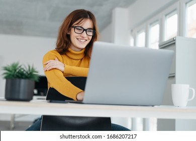 Young businesswoman smiling as she works on a laptop seated at a table in an office in a low angle close up view