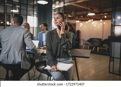 Young businesswoman smiling and talking on her cellphone while sitting in an office with colleagues in the background - Shutterstock ID 1722971407