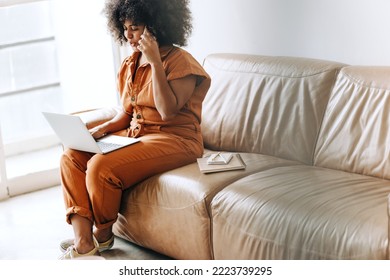 Young Businesswoman Speaking On The Phone While Working On A Laptop In An Office Lobby. Black Businesswoman Communicating With Her Business Partners Over A Phone Call.