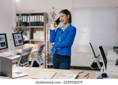 Young businesswoman standing in modern office using smartphone, smiling while talking on mobile phone, computers with data analysis charts on screens in background - Powered by Shutterstock