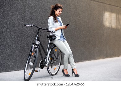 Young businesswoman standing on a city street with bicycle looking at phone. - Powered by Shutterstock