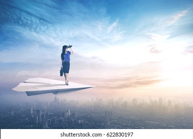 Young businesswoman standing on a paper aircraft and flying above a city while looking through binoculars