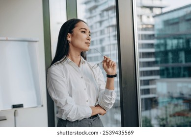 Young businesswoman stands by office window gazing thoughtfully at city skyline - Powered by Shutterstock