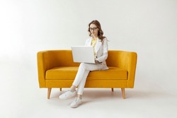 Young Businesswoman In Suit Sits On Comfortable Soft Sofa And Uses Laptop, Girl In Formal Wear Is Typing On Computer On Yellow Couch On White Isolated Background