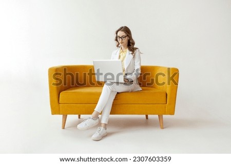 young businesswoman in suit sits on comfortable soft sofa and uses laptop, girl in formal wear is typing on computer on yellow couch on white isolated background