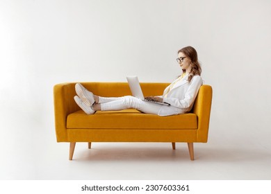 young businesswoman in suit uses laptop on comfortable soft sofa, girl in formal wear is typing on computer on yellow couch on white isolated background