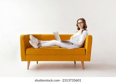 young businesswoman in suit uses laptop on comfortable soft sofa, girl in formal wear is typing on computer on yellow couch on white isolated background