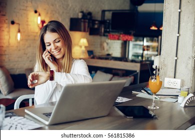 Young Businesswoman Talking On The Phone And Checking The Time Beside Laptop On Table In Cafe