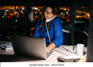 Young businesswoman talking on the phone and using a laptop at her office desk late into the night in front of windows overlooking the city  - Powered by Shutterstock