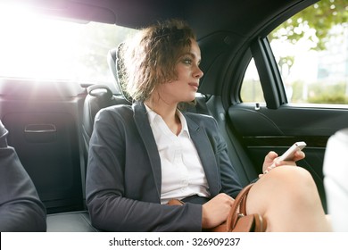 Young Businesswoman Traveling To Work In The Luxury Car On The Back Seat Holding Mobile Phone And Looking Outside Window.