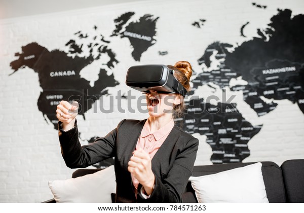 Young businesswoman trying\
virtual reality glasses sitting indoors with world map on the\
background