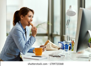 Young businesswoman using cell phone while eating a cookie at her office desk. 