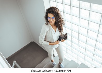Young businesswoman using a digital tablet while walking up a staircase in an office. Businesswoman ascending office staircase. Professional Businesswoman Walking on Stairs in an Office Lobby 