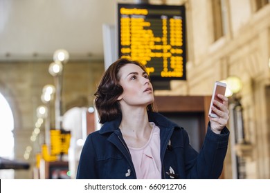 Young Businesswoman Is Using Her Smart Phone To Check For Her Train Time On The Departure Board. 