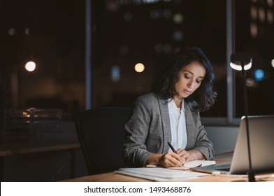 Young businesswoman using a laptop and writing notes while working overtime at her office desk late in the evening - Powered by Shutterstock