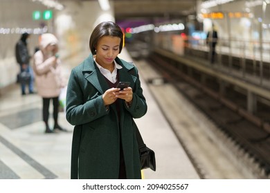 Young Businesswoman Waiting On Subway Platform For Carriage Reading Text Or Business Email On Smartphone. Busy African Woman With Mobile Phone Chatting With Colleague While Returning Home From Office