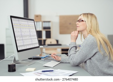 Young Businesswoman Wearing Glasses Sitting At Her Desk In The Office Working On A Desktop Computer, Side View