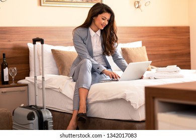 Young Businesswoman Working From Hotel Room On Business Trip, Woman Sitting On Bed And Using Laptop.