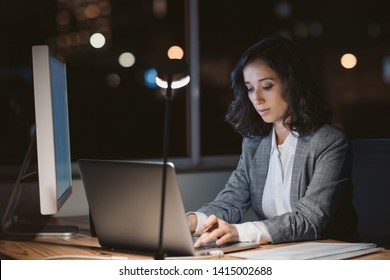 Young businesswoman working overtime alone at her desk in an office late at night with city lights glowing in the background - Shutterstock ID 1415002688