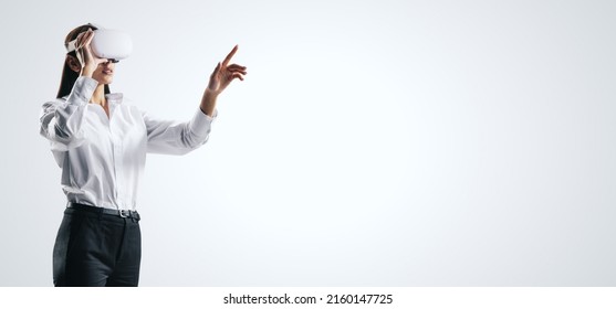 Young businesswoman working in virtual reality environment in VR glasses, presses by her finger on virtual interface on abstract white backdrop. Innovate and future technology concept, mock up - Powered by Shutterstock