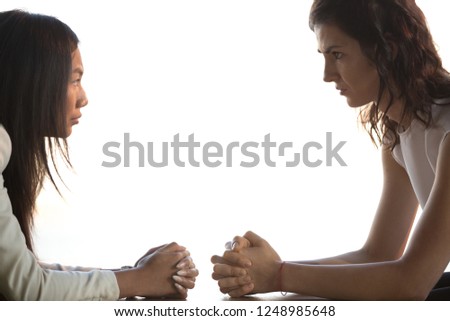 Young businesswomen sitting at desk with hands clenched together staring at each other unkindly, side view. Women struggling for leadership, having different opinions or discrimination at work concept