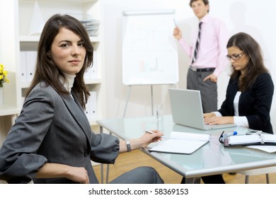 Young businesswomen sitting at desk, writing notes. Businesswoman standing in background, drawing chart on whiteboard. Selective focus on woman in front.