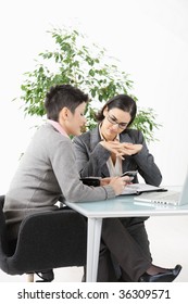 Young Businesswomen Sitting At Office Desk, Looking At Smart Phone.