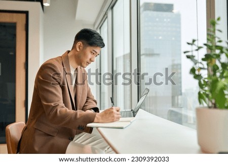 Young busy Asian business man manager working on laptop in office writing notes. Professional businessman analyst using computer checking corporate data, watching online webinar, elearning concept.