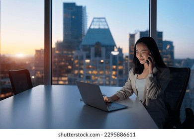 Young busy Asian business woman executive working on laptop making call at night in dark corporate office. Professional businesswoman manager talking to client using computer, city evening window view