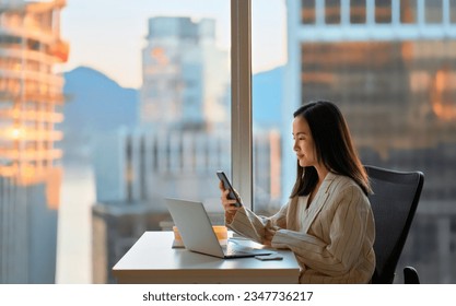 Young busy Asian business woman leader holding cellphone device using mobile phone, looking at smartphone checking financial market app working in modern corporate office with big window. - Powered by Shutterstock