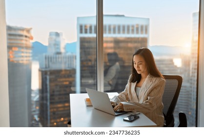 Young busy Asian business woman executive working on laptop in corporate office. Professional businesswoman marketing sales manager using computer technology sitting at table, city view from window. - Powered by Shutterstock