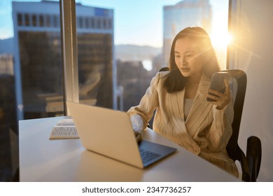 Young busy Asian business woman leader holding mobile phone, looking at laptop computer working with technology devices working in modern corporate office lit with sun light through big window. - Powered by Shutterstock