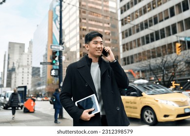 Young busy happy Asian business man office professional holding cellphone in hands walking on big city urban street making corporate business call, talking on cellular phone standing in downtown.