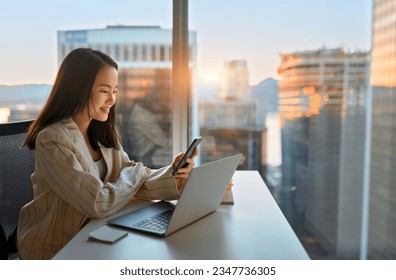 Young busy happy Asian business woman manager using mobile cell phone tech in office. Professional female executive holding smartphone, working, checking cellphone sitting at desk at big window. - Powered by Shutterstock