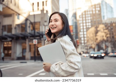 Young busy happy Asian business woman office professional holding cellphone in hands walking on big city urban street making corporate business call, talking on the cellular phone. Authentic shot