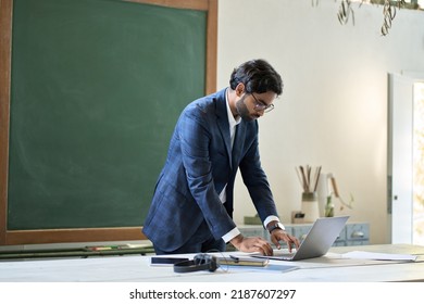 Young busy indian businessman working on laptop standing in office at work desk. Arabic teacher professor using computer for online education, remote teaching classes in front of blackboard. - Shutterstock ID 2187607297