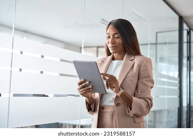 Young busy professional African American business woman company manager sales executive wearing suit holding tab using digital tablet computer standing in office managing financial data. - Powered by Shutterstock