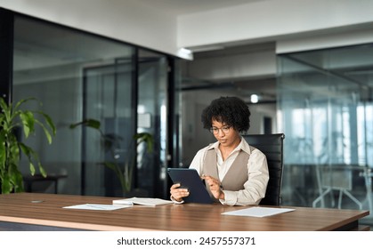 Young busy professional African American business woman company executive or entrepreneur using digital tablet working sitting at desk in office looking at tab, managing financial project data. - Powered by Shutterstock