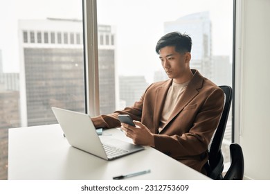 Young busy serious Asian Japanese business man worker executive holding cellphone using mobile phone looking at laptop checking financial market online in app working in corporate office at desk. - Powered by Shutterstock