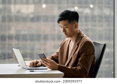 Young busy smart serious Asian Japanese business man executive holding cellphone device using mobile cell phone looking at smartphone working in corporate office with laptop computer technology.