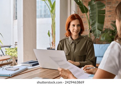 Young busy smiling professional business woman with red hair lawyer, financial advisor or company manager consulting female client, talking to colleague working in office using laptop sitting at desk. - Powered by Shutterstock