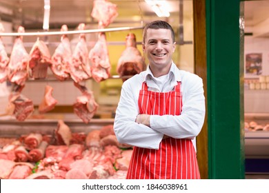 A young butcher in a red apron smiling at the camera and standing in front of butcher shop window.