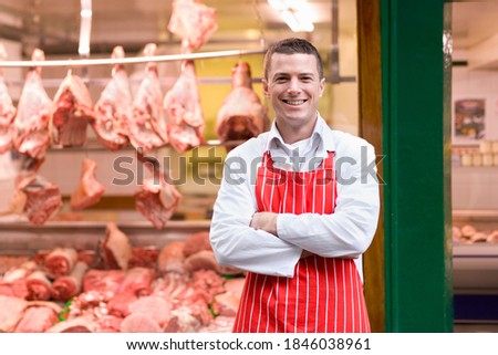 A young butcher in a red apron smiling at the camera and standing in front of butcher shop window.