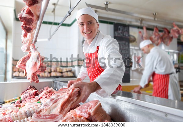 A young butcher smiling and holding meat near the\
camera in a meat shop.